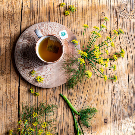 A cup of Fennel Tea rests on a wooden table, encircled by yellow wildflowers and green plant stems, capturing the spirit of herbalism and supporting healthy digestion.