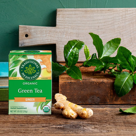 Box of Green Tea Ginger with fresh ginger root and vibrant green leaves on a wooden surface.