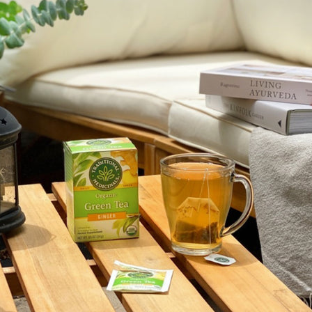 A cup of Green Tea Ginger on a wooden table with tea bags and books nearby on a cozy patio.