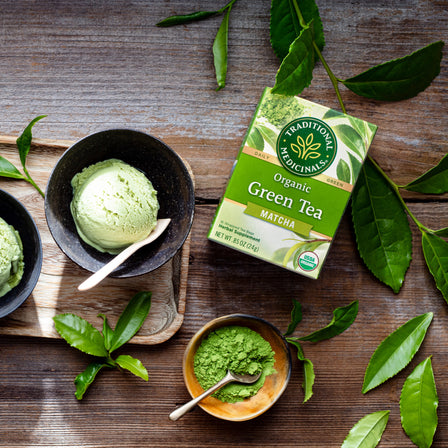 Green Tea Matcha ice cream, a green tea box, and organic Green Tea Matcha powder grace a wooden table adorned with scattered tea leaves, evoking the traditional essence of Japan.