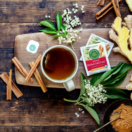 A cup of Herbal Cold Care™ Tea rests on a wooden board, accompanied by ginger, cinnamon sticks, and flowers, providing soothing comfort.