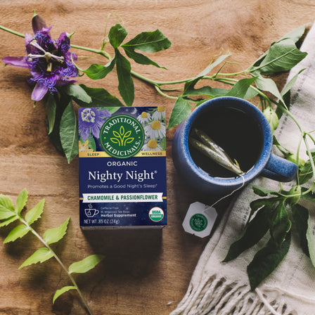 Box of Nighty Night® Tea showcasing calming chamomile and soothing passionflower, accompanied by a cup of tea placed on a cloth and encircled by green leaves. Ideal for promoting a restful sleep.