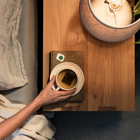 A hand gently cradles a mug of Nighty Night® Tea, resting on a wooden table alongside a softly glowing lamp and a packet of tea bags.