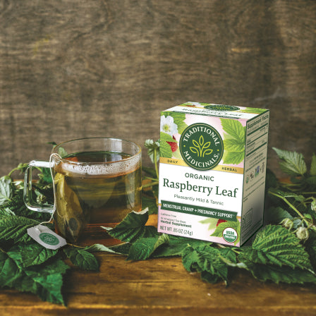 A glass cup of herbal tea sits beside a box of Raspberry Leaf Tea on a wooden table surrounded by green leaves, offering natural support for menstrual cramps.