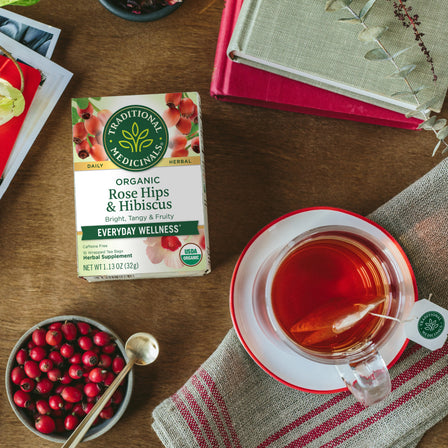 A box of Rose Hips & Hibiscus Tea and a cup sit elegantly on the table, surrounded by cranberries, a spoon, and a stack of books. The soothing aroma hints at organic tea infused with delicate rose hips.