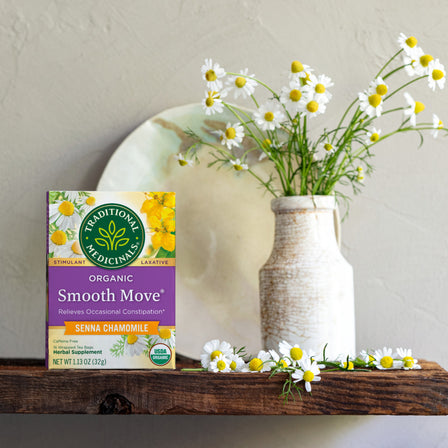 A box of Smooth Move® Chamomile Tea rests on a wooden shelf, promising a gentle overnight remedy with a vase of blooming chamomile flowers in the background. It's perfect for soothing relaxation and natural constipation relief.