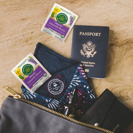 A Smooth Move® Tea pouch with senna tea packets, a passport, and a patterned cloth sits on the wooden surface, promising constipation relief for your journey.