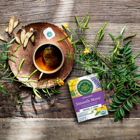 A cup of senna tea sits next to an opened box of Smooth Move® Tea on a rustic wooden table, surrounded by leaves and roots, offering gentle overnight relief.