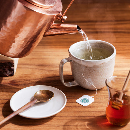 A copper kettle pours hot water into a mug containing an organic tea bag of Spearmint Tea. Next to it, honey in a jar and a spoon are placed on the wooden table, creating the perfect setup for a refreshing beverage.