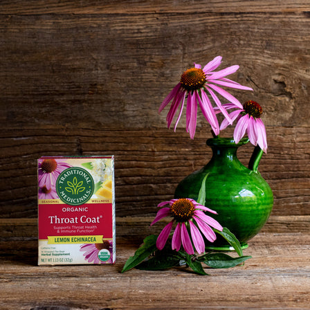 A box of Throat Coat® Lemon Echinacea Tea sits beside a green vase adorned with pink flowers on a wooden backdrop, making it ideal for enhancing throat health.