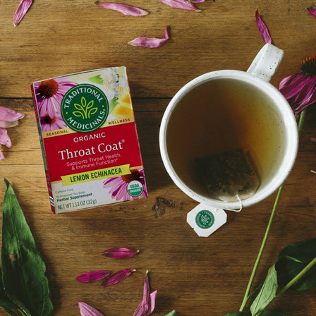 A box of Throat Coat® Lemon Echinacea Tea is placed next to a steaming mug, surrounded by flower petals on a wooden surface. Ideal for promoting throat health and supporting your immune system.