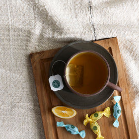 Enjoy a cup of peppermint tea on a wooden tray with a lemon slice and Throat Coat® Eucalyptus Mint Lozenges, 30ct on a textured cloth—perfect for soothing sore throats.