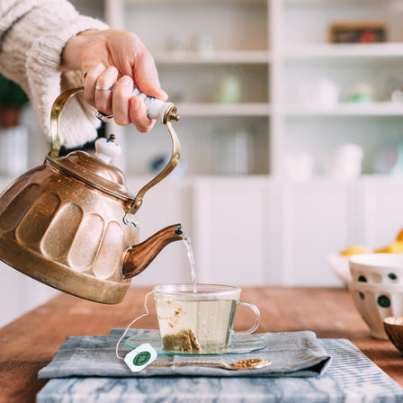 A hand pours hot water from a copper kettle into a glass teacup on a table, releasing the soothing aroma of Turmeric & Ginger with Meadowsweet Tea as the tea bag unfurls.